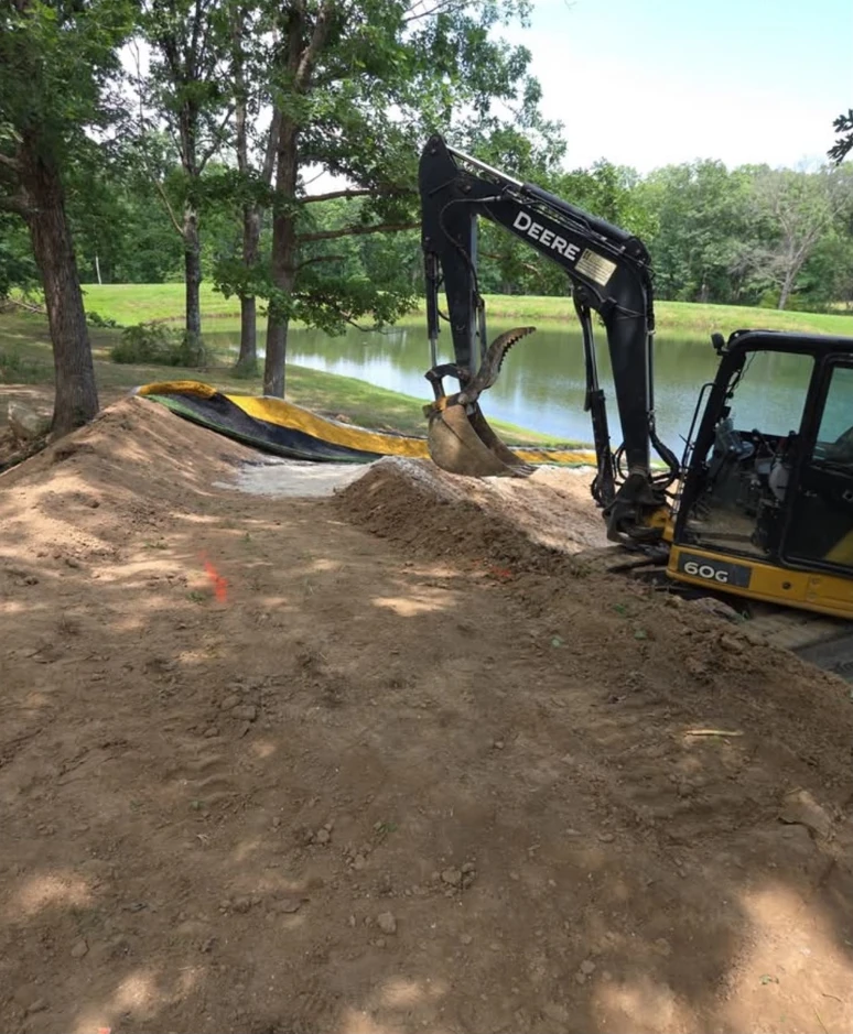 An excavator working on a water-slide project beside a pond and trees.